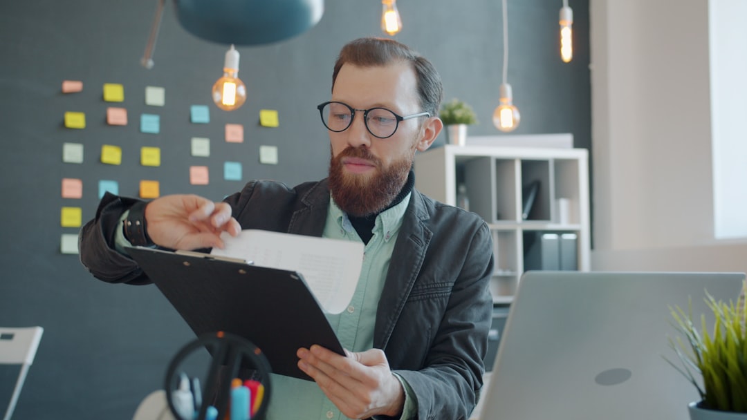 Male office worker is reading papers and using laptop checking information working at desk alone. Businesspeople, job and modern technology concept.